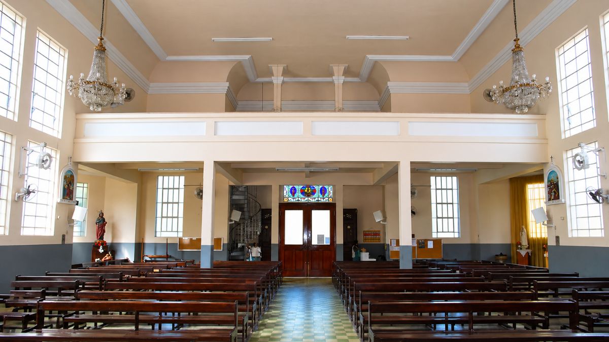 Fotografia frontal centralizada de uma igreja cinza com torre sineira escalonada, relógio, porta de madeira e escadaria de acesso.
