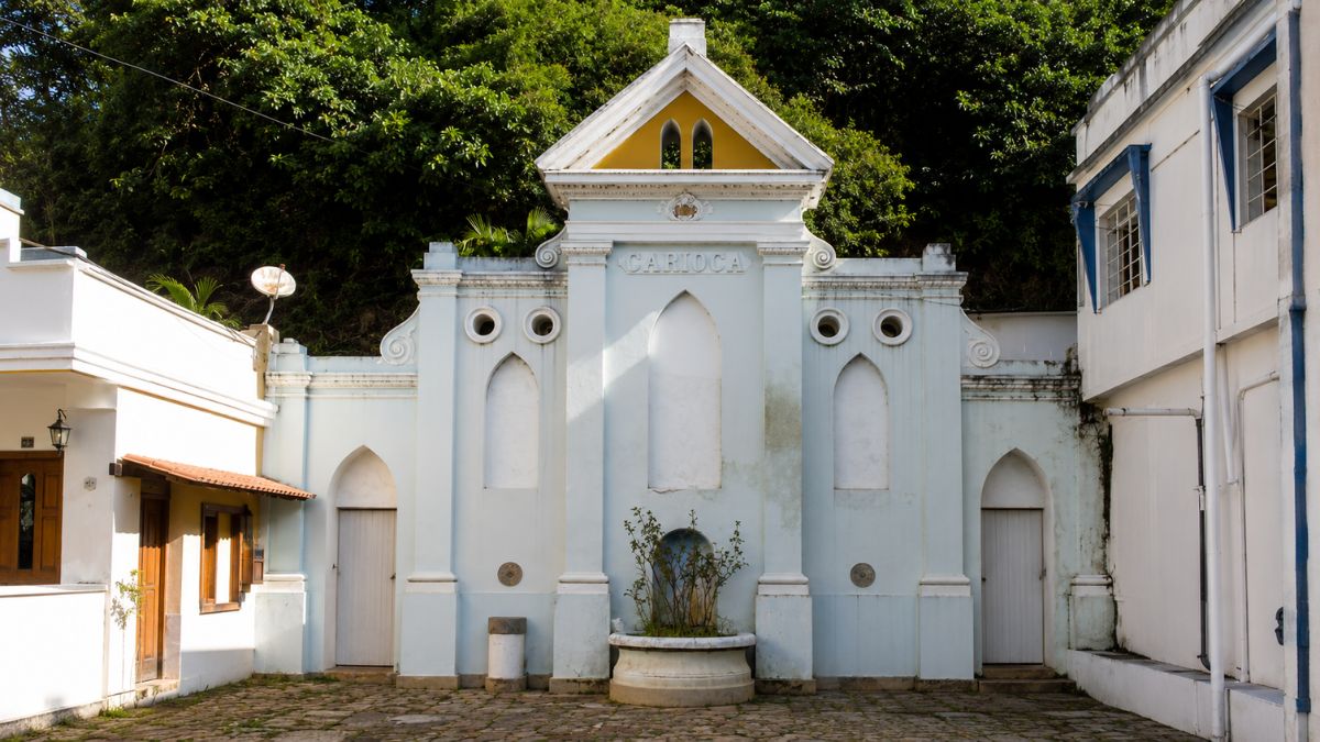 Fotografia central da fachada azul claro de um chafariz com frontão triangular amarelo, nichos em arco e portas laterais brancas.