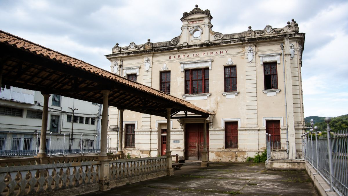 Edifício histórico de dois pavimentos com frontão decorado e uma cobertura externa de telhas coloniais sobre colunas de sustentação.
