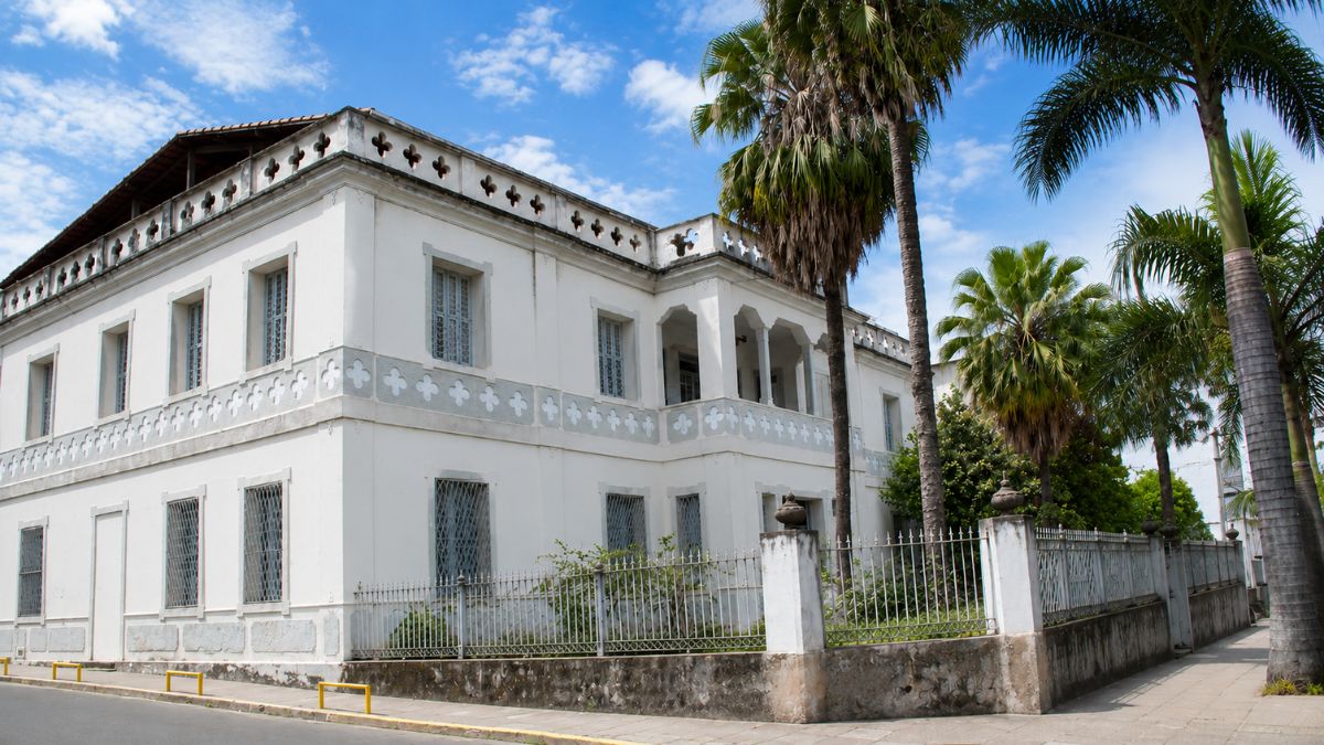 Edifício histórico de dois pavimentos na cor branca com platibandas ornamentadas por quadrifólios góticos ladeado por palmeiras sob céu azul.