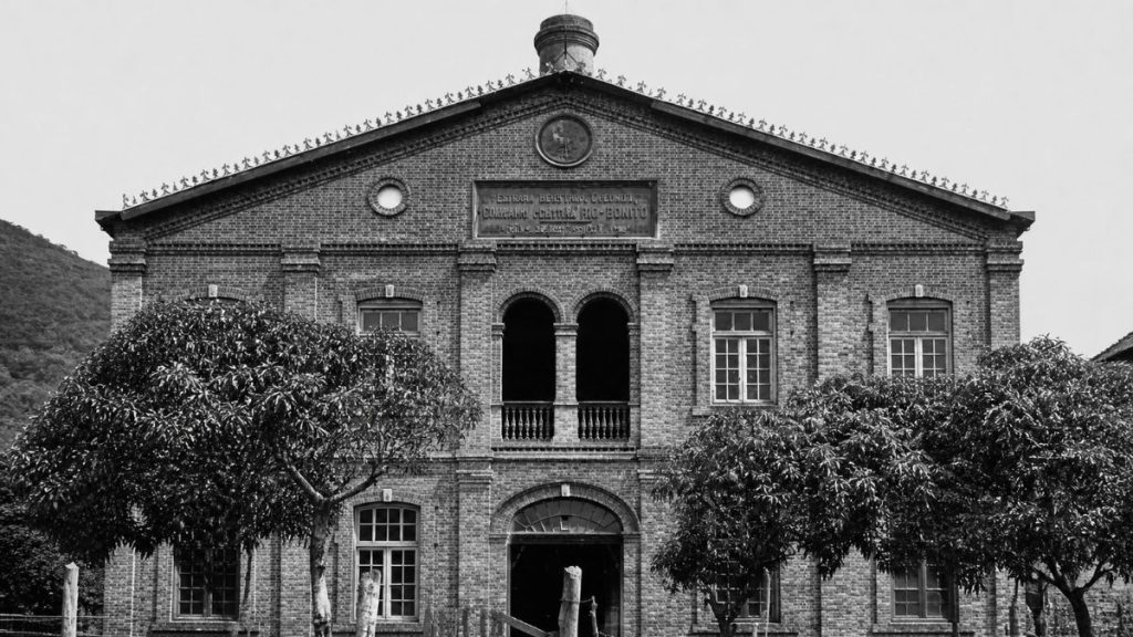 Fotografia antiga frontal de um prédio de dois andares em tijolos com grande frontão triangular e relógio circular central.