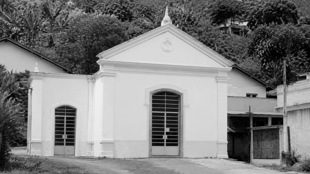 Fotografia em preto e branco de uma capela neoclássica com frontão triangular e cruz centralizada situada diante de uma encosta.