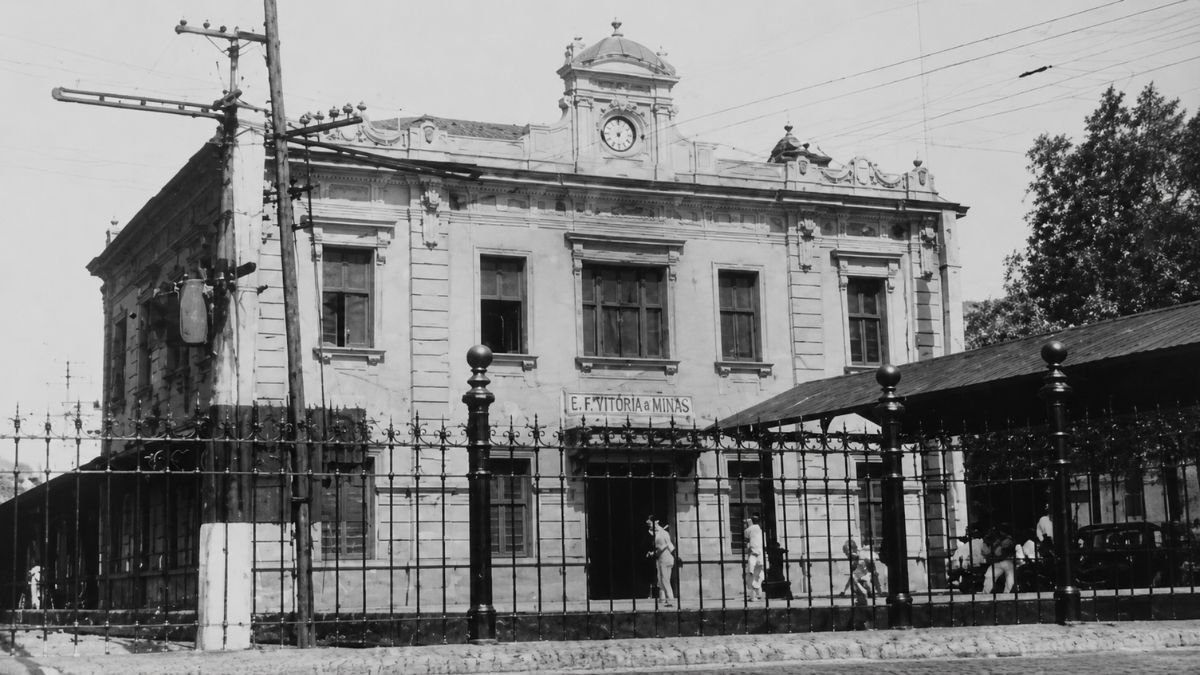 Fotografia em preto e branco de prédio histórico com relógio central e gradil metálico.