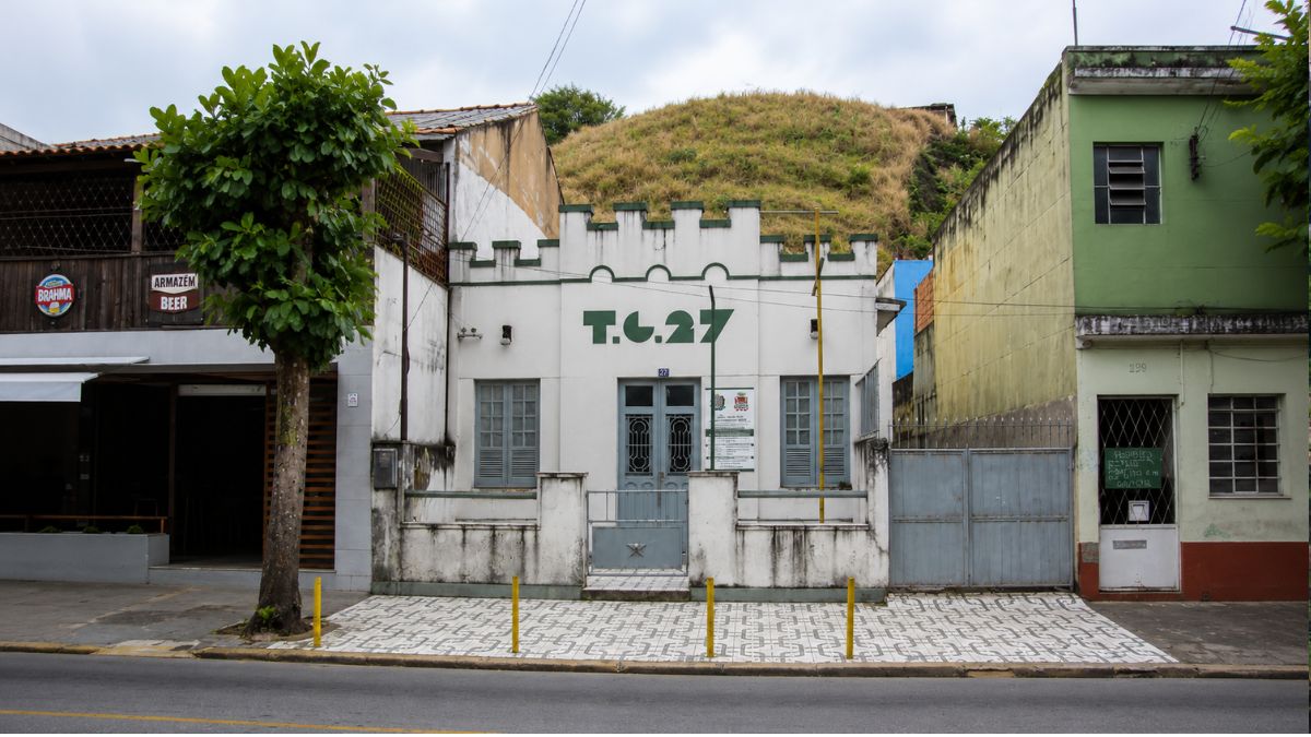 Fachada de uma casa térrea branca com a inscrição TG 27 em verde, possuindo platibanda em formato de ameias militares.