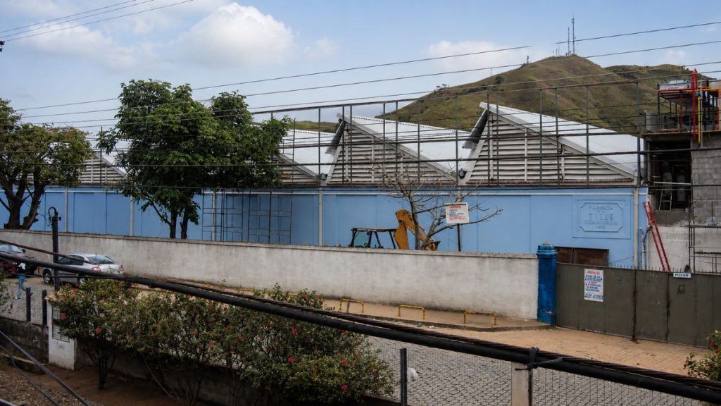 Fotografia em nível da rua mostrando muro branco, fachada azul com telhado serrilhado, andaimes de obra e morros ao fundo.