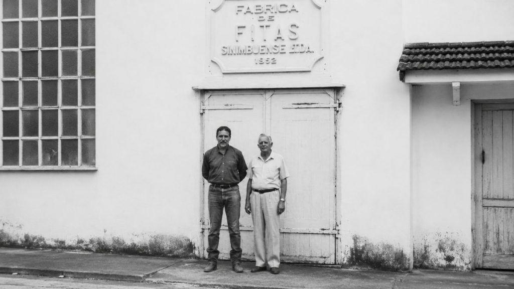 Registro antigo de dois homens de pé em frente a um portão de madeira sob o letreiro da fábrica histórica.