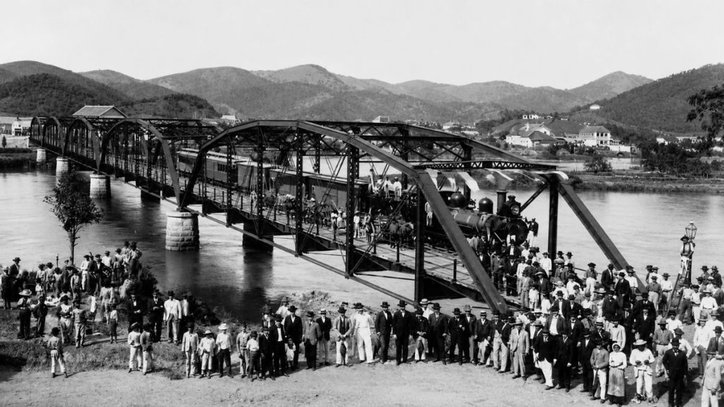 Fotografia histórica em preto e branco de uma locomotiva sobre a ponte metálica com centenas de pessoas reunidas na margem.