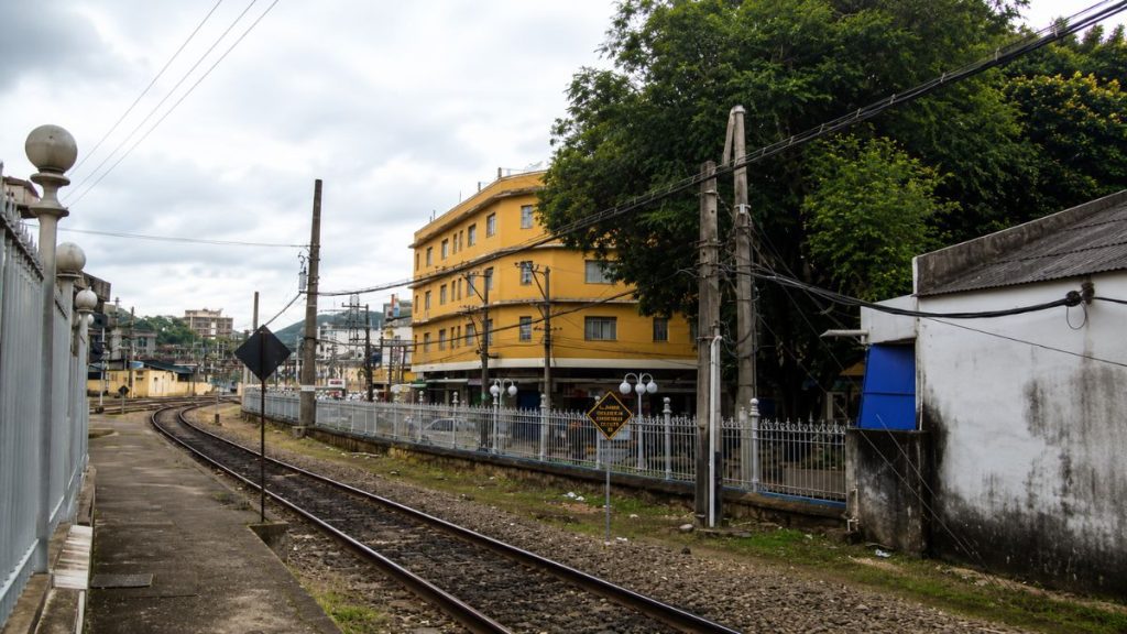 Trilhos de trem em curva acompanhados por uma plataforma de embarque e cercas metálicas com prédios amarelos ao fundo.