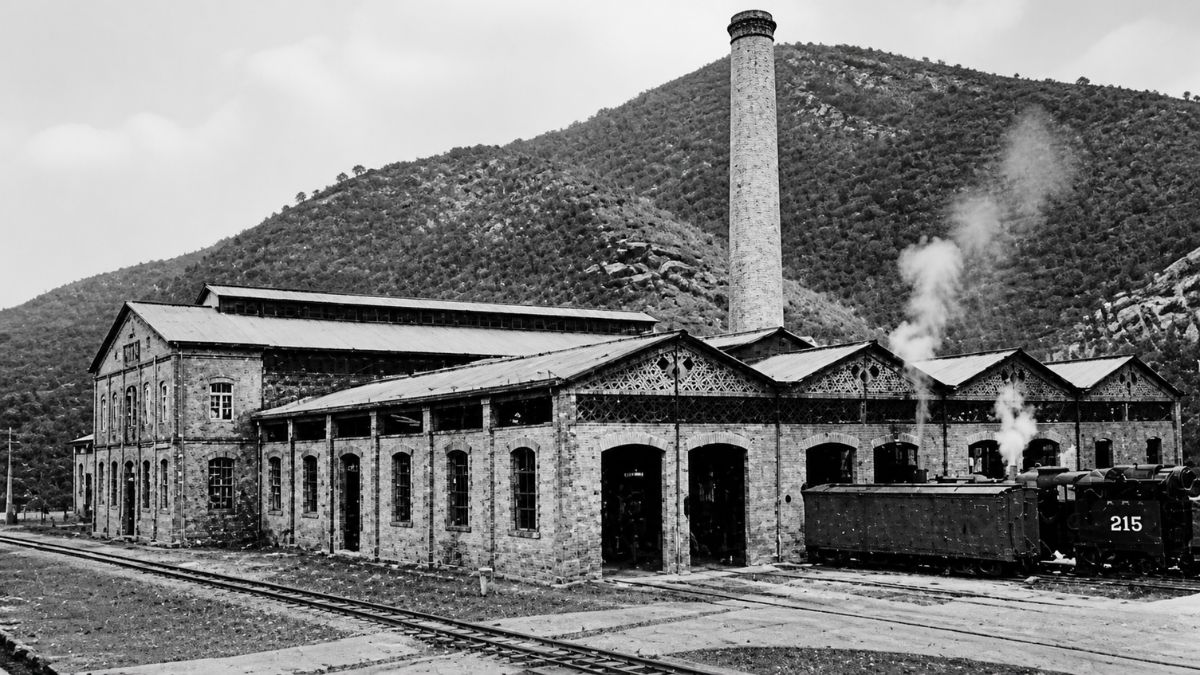 Fotografia em preto e branco de oficina ferroviária com chaminé alta e locomotiva soltando fumaça.