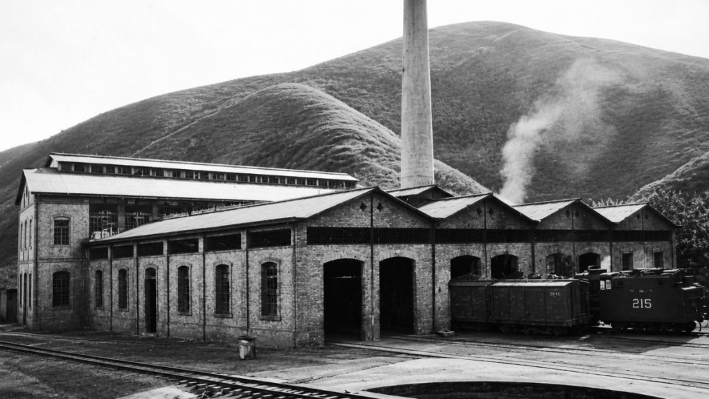 Fotografia antiga em preto e branco de galpões industriais com trilhos, locomotiva numerada duzentos e quinze e chaminé ao fundo.