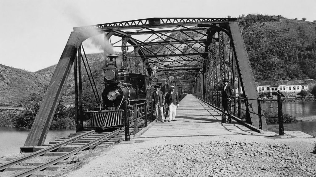 Fotografia histórica em preto e branco de uma locomotiva a vapor sobre trilhos na ponte metálica com três homens posando.