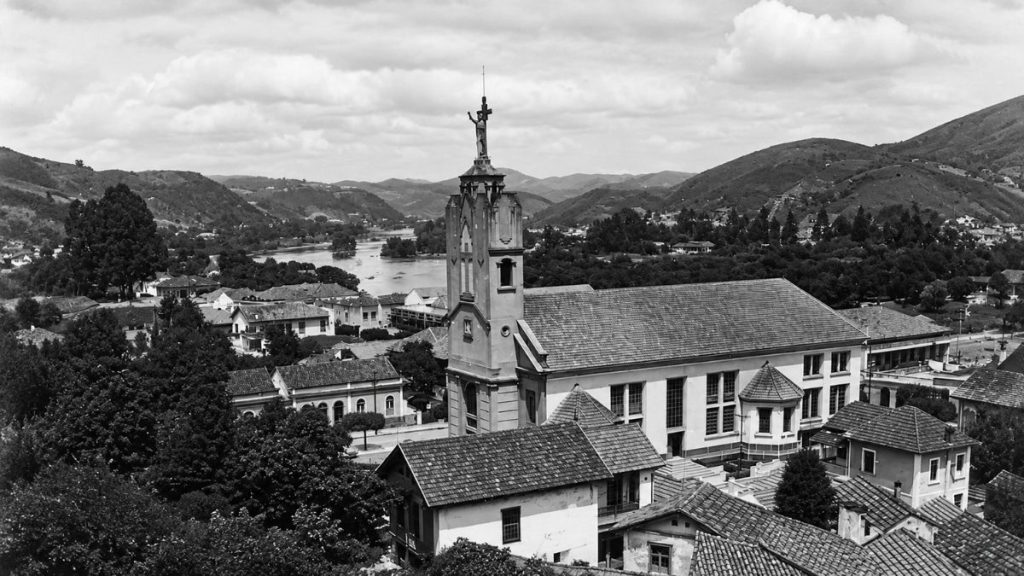 Fotografia panorâmica antiga em preto e branco mostrando a igreja, o casario do entorno, o rio e montanhas ao fundo.