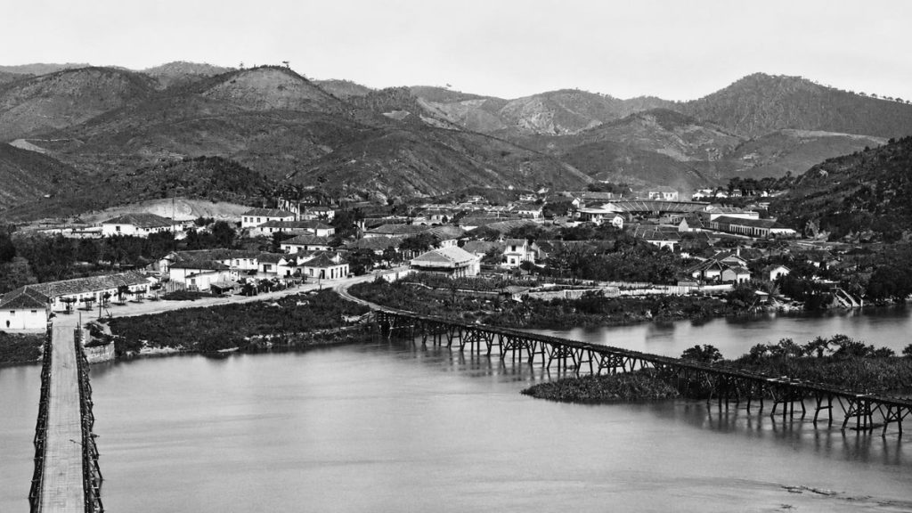 Fotografia antiga em preto e branco de uma cidade histórica com uma longa ponte de madeira sobre o rio largo.