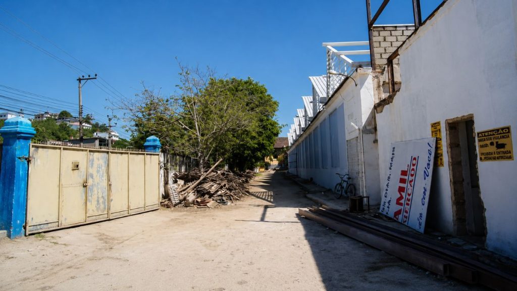 Fotografia do corredor externo da fábrica com entulhos acumulados, paredes brancas, portão de ferro bege e céu azul limpo.