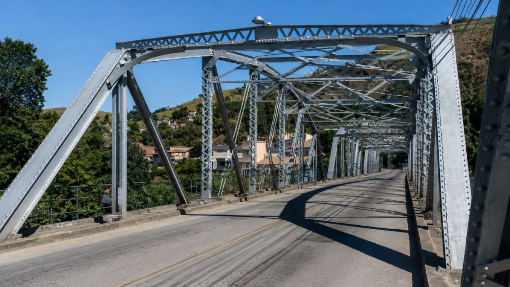 Vista frontal da pista asfáltica da Ponte Getúlio Vargas emoldurada por treliças metálicas cinzas e montanhas verdes ao fundo.