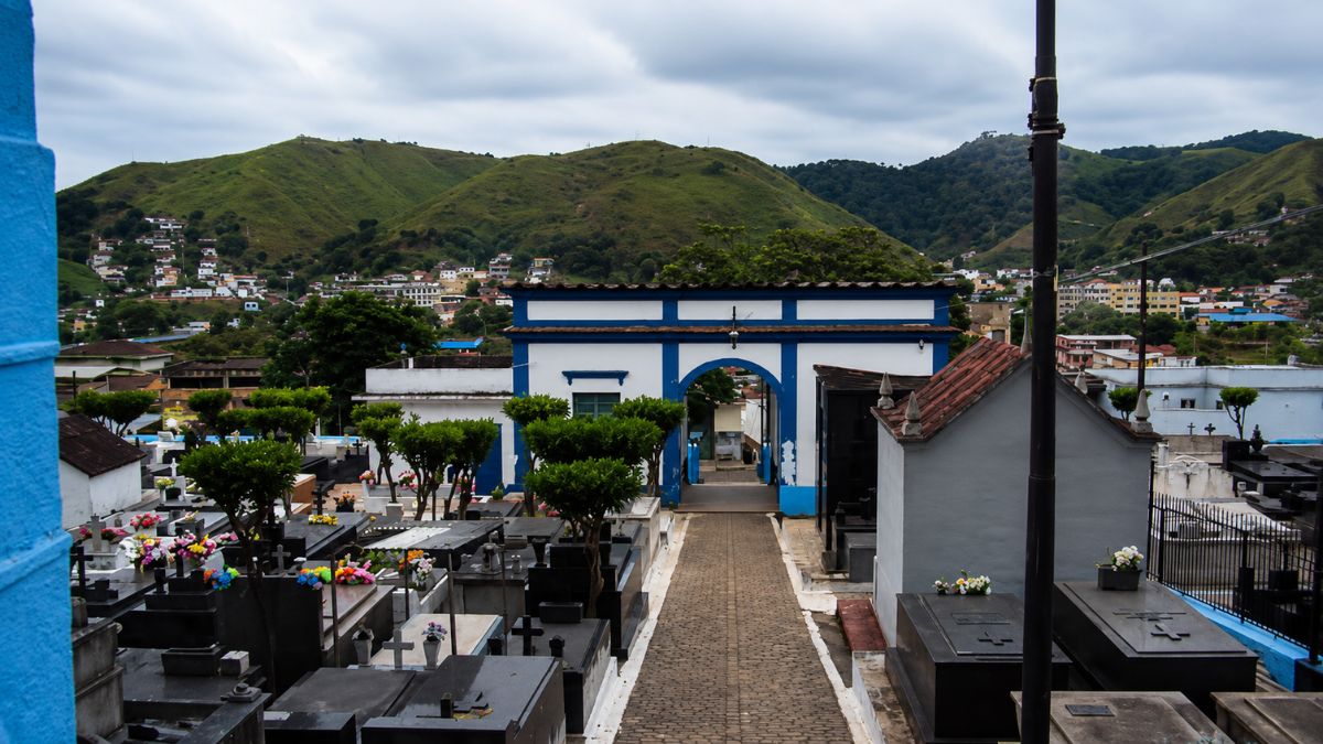 Fotografia em ângulo elevado mostrando o caminho central de paralelepípedos, túmulos de granito e o pórtico azul e branco com montanhas ao fundo.