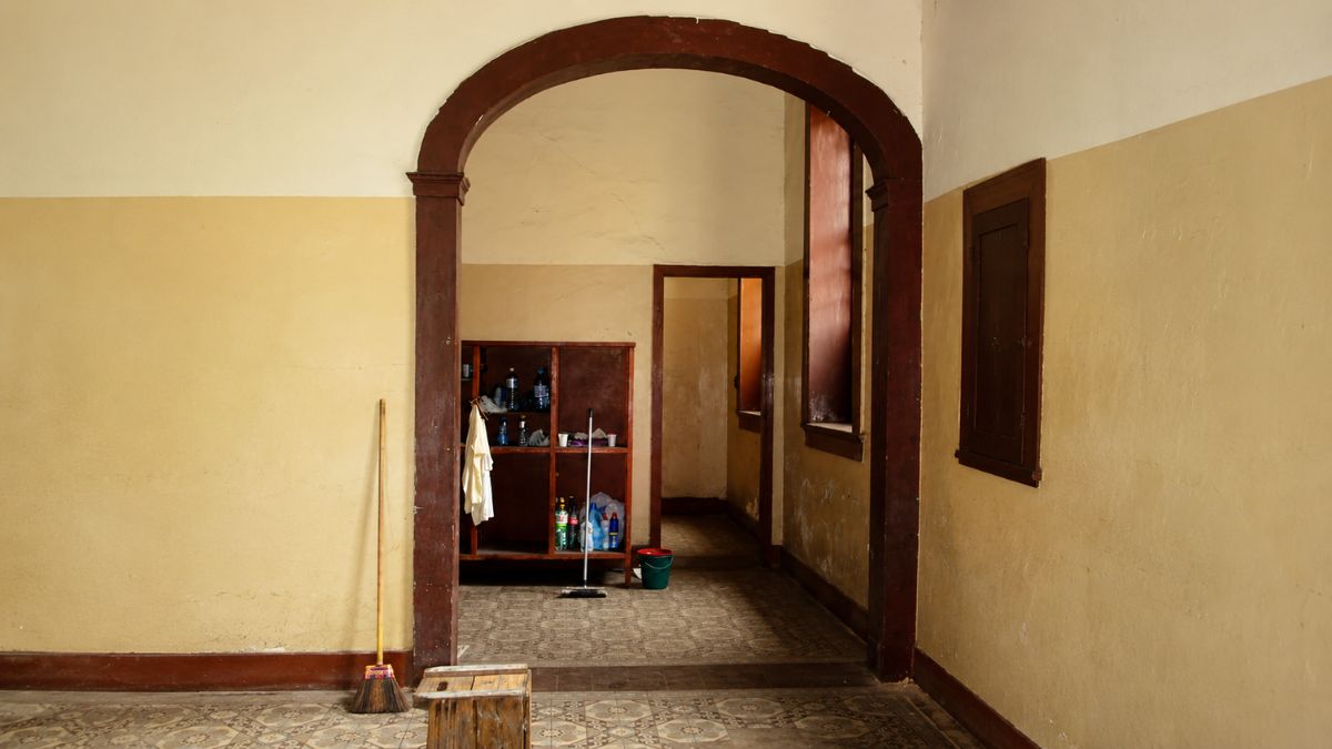 Vista interna de um corredor através de um grande arco de madeira escura com piso decorado e paredes em bege.