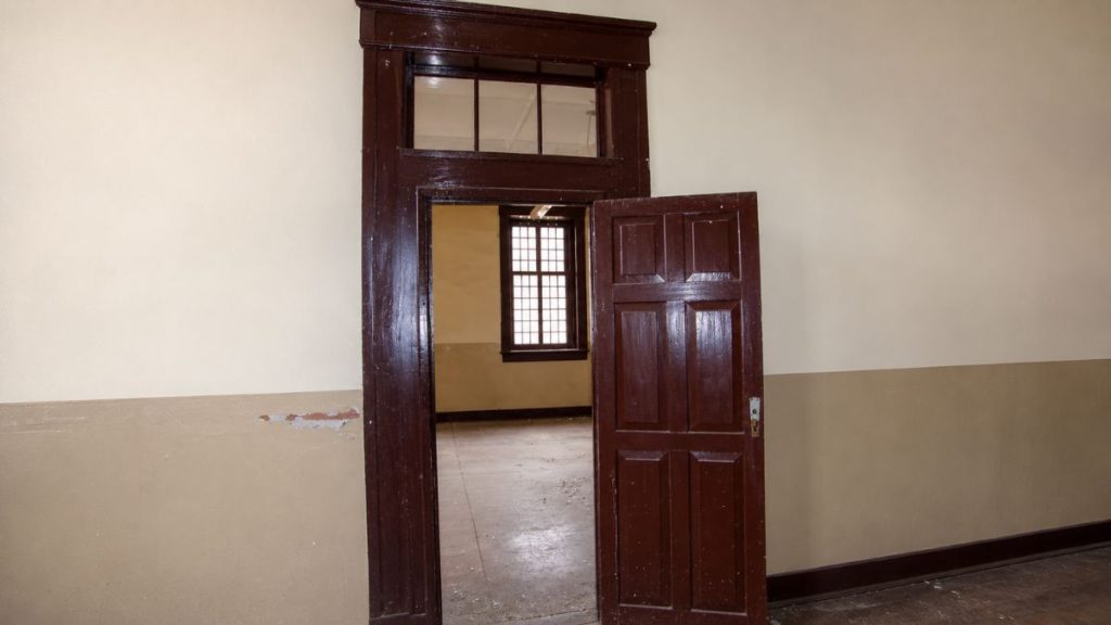 Vista de uma porta de madeira escura aberta revelando o interior de uma sala vazia com janela ao fundo.