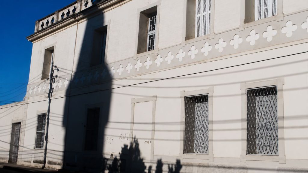 Fachada lateral de prédio antigo com janelas protegidas por grades e friso decorativo sob sombra projetada de poste elétrico.
