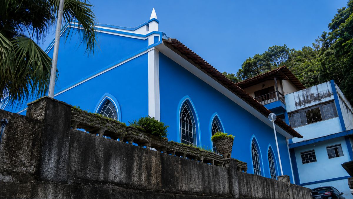 Foto em ângulo baixo da lateral de uma igreja azul com janelas em arco ogival. Um muro de pedra rústico está em primeiro plano e um edifício de apoio aparece ao fundo.
