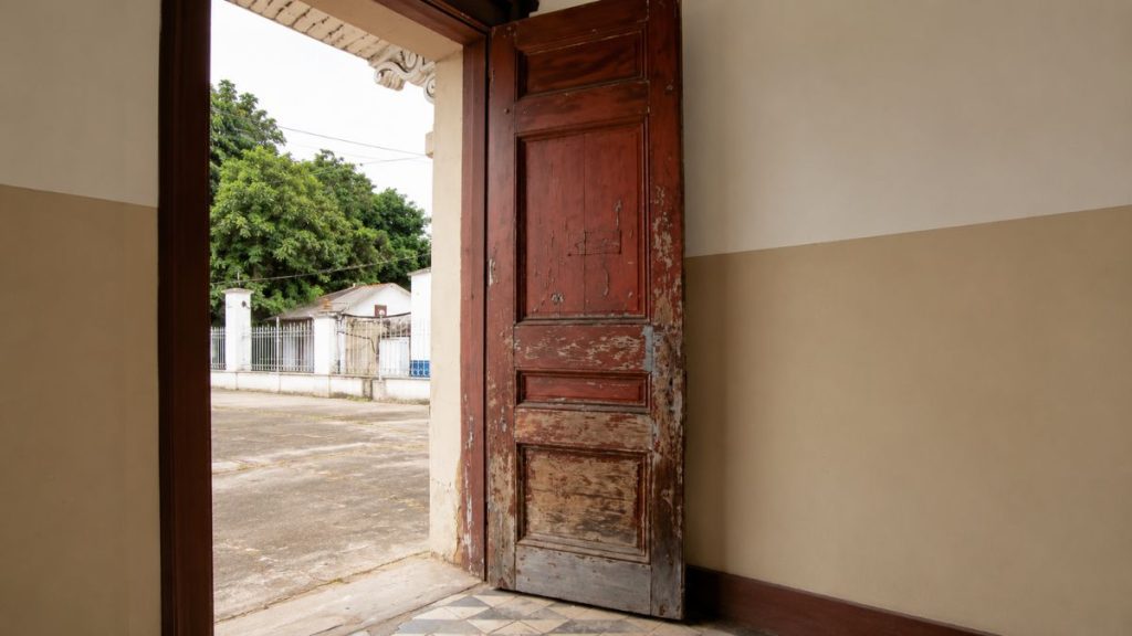 Vista de dentro para fora através de uma porta de madeira antiga aberta revelando um pátio externo e árvores.