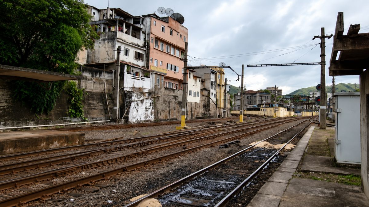 Trilhos ferroviários em primeiro plano com edifícios residenciais construídos sobre um muro de contenção ao lado da via férrea.
