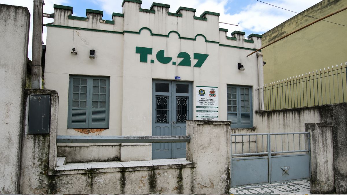 Fachada frontal centralizada do prédio TG 27 com platibanda em ameias, portão de ferro cinza e placa da Junta Militar.