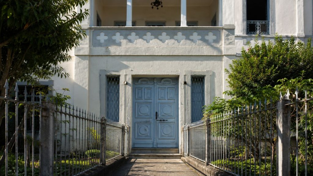 Close da entrada principal com porta azul almofadada ladeada por janelas estreitas e caminho isolado por gradis de ferro cinza.
