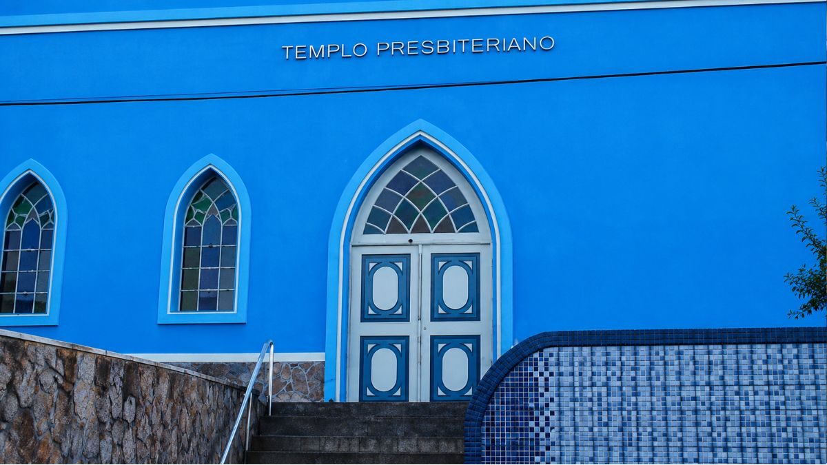 Vista frontal da entrada de uma igreja azul com a inscrição Templo Presbiteriano na fachada. O portal central e as janelas possuem arcos ogivais. Muros de pedra e azulejos emolduram a escada.