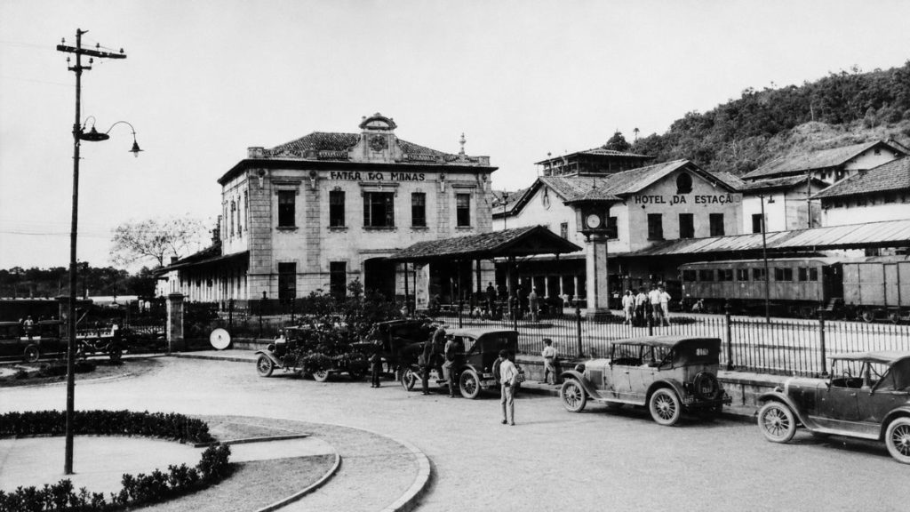 Fotografia antiga em preto e branco de uma estação ferroviária com carros de época estacionados e pessoas na plataforma.