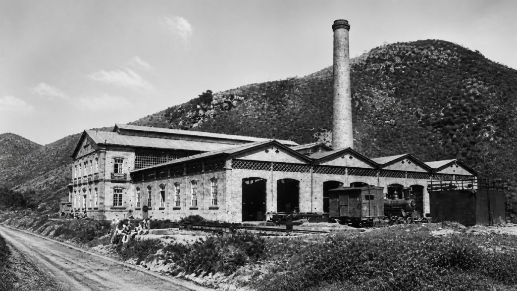 Fotografia antiga em preto e branco mostrando o complexo industrial do engenho com locomotiva e alta chaminé sob montanha.