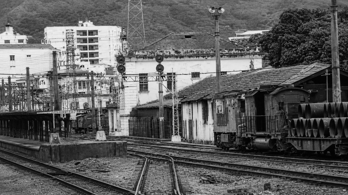 Fotografia em preto e branco mostrando trilhos de trem, vagões de carga e estação.