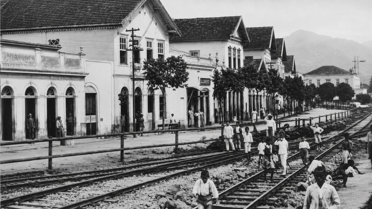 Fotografia antiga em preto e branco mostrando prédios coloniais e pessoas sobre trilhos.
