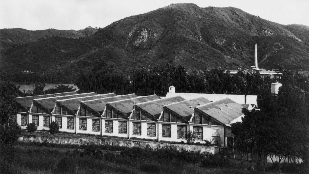 Fotografia antiga em tons de cinza mostrando o galpão industrial com telhado serrilhado em meio a vegetação e morros.