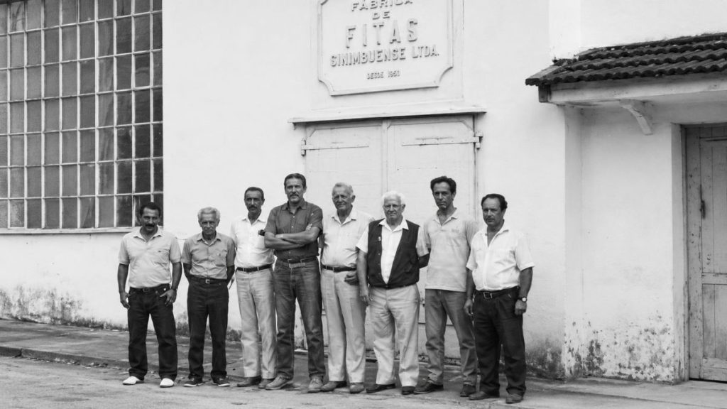 Fotografia antiga de oito homens perfilados diante de uma parede branca com letreiro da Fábrica de Fitas Sinimbuense Ltda.