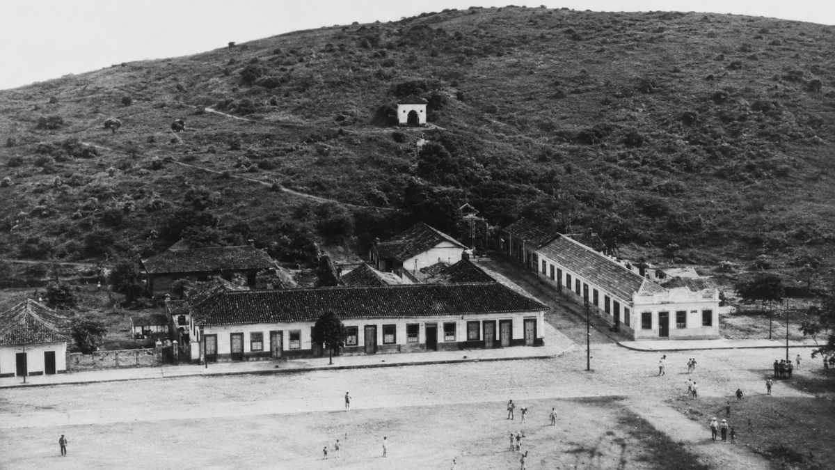 Fotografia aérea antiga em preto e branco mostrando casas coloniais, ruas de terra e o chafariz com a caixa d’água na encosta.