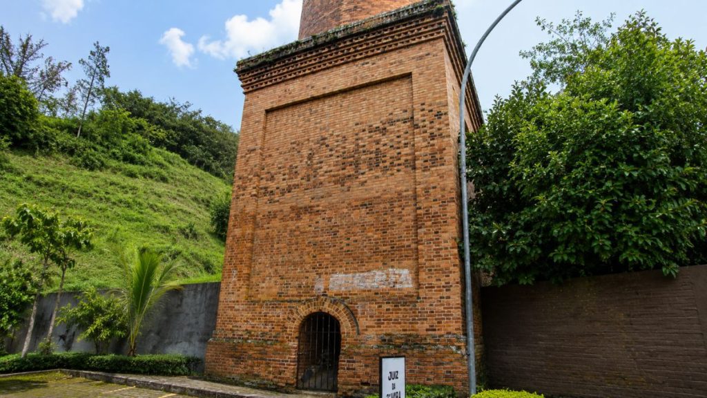 Base quadrada de tijolos aparentes com portão de ferro em arco pleno e frisos decorativos sob céu com nuvens.
