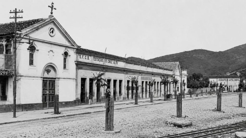 Fotografia antiga em preto e branco de uma estação ferroviária de fachada longa com uma pequena capela branca à esquerda.