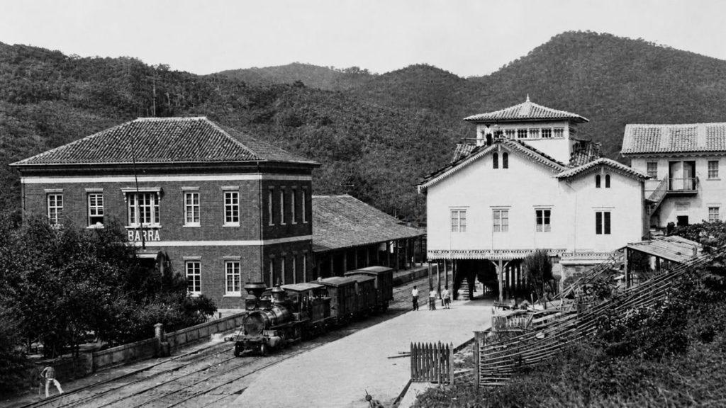 Fotografia antiga em preto e branco de 1880 mostrando uma locomotiva a vapor entre prédios históricos e montanhas ao fundo.