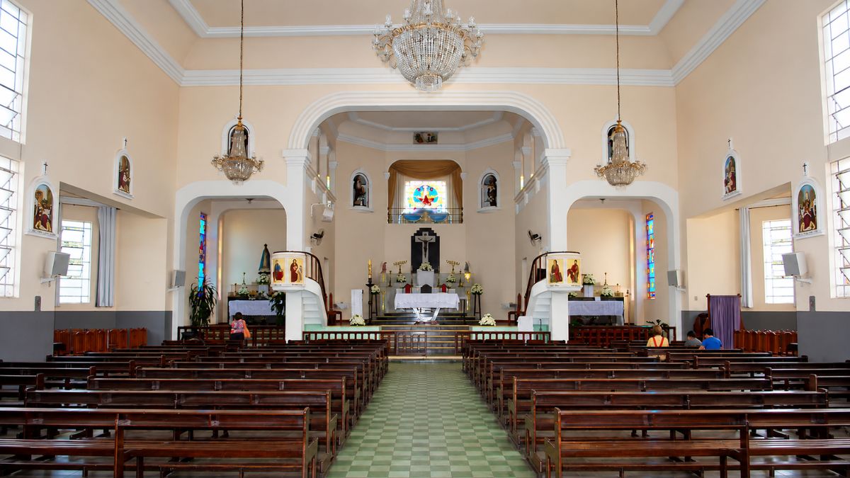 Fotografia interna da nave central mostrando bancos de madeira, teto com candelabros de cristal e altar-mor ao fundo com crucifixo.