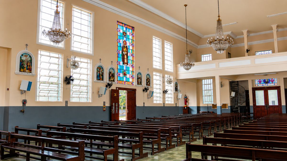 Fotografia interna da nave mostrando bancos de madeira, paredes claras com pinturas sacras, lustres de cristal e grande vitral colorido.