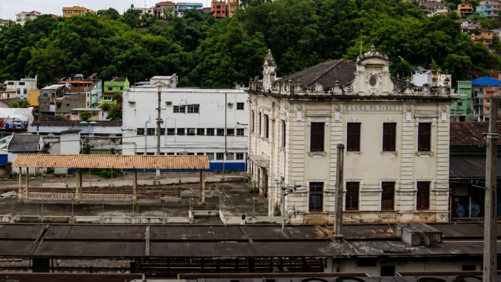 Fotografia lateral do edifício neoclássico da estação ferroviária com telhados de plataformas em primeiro plano e morros arborizados ao fundo.