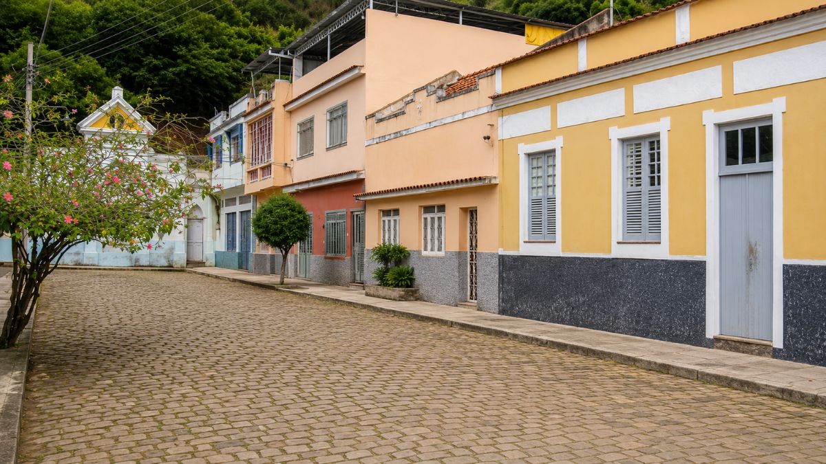 Fotografia em ângulo lateral mostrando uma rua de paralelepípedos com casas coloridas e o chafariz histórico ao fundo sob a colina.