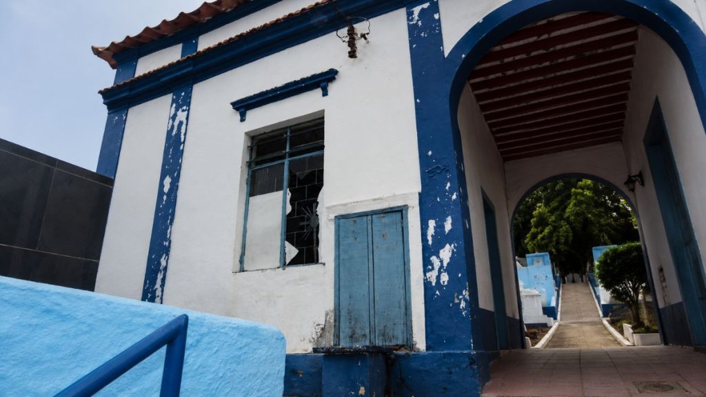 Fotografia de um ângulo inclinado mostrando a fachada lateral branca e azul do pórtico com uma janela de vidro quebrado.