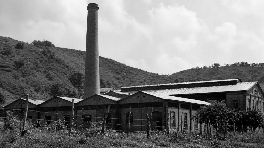 Fotografia antiga em preto e branco de galpões industriais de tijolos com telhados em shed e alta chaminé cilíndrica.