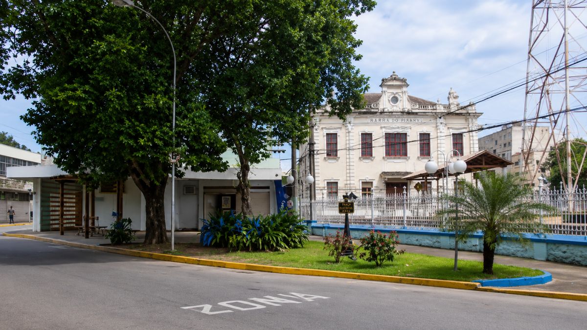 Rua asfáltica em curva com canteiros arborizados em primeiro plano e a fachada da estação ferroviária neoclássica ao fundo.