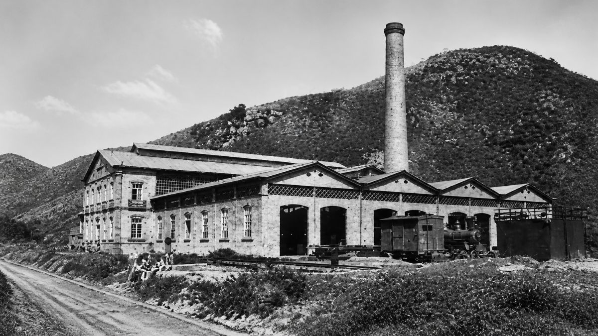 Fotografia em preto e branco de complexo industrial com chaminé alta diante de morro pedregoso.