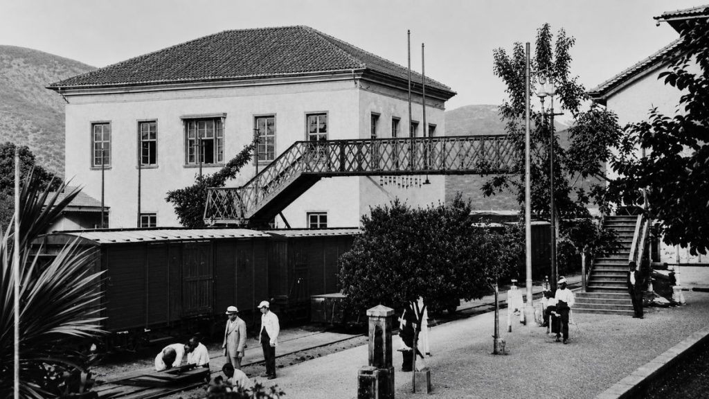 Fotografia antiga em preto e branco de 1880 mostrando uma passarela metálica sobre trilhos com vagões de carga e pessoas.