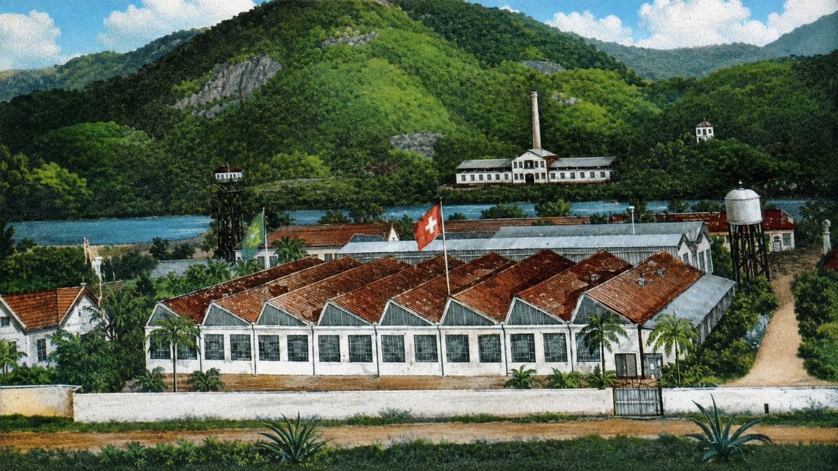 Fotografia colorizada da antiga fábrica com telhado em formato de serra, muros brancos, vegetação densa e montanhas ao fundo.