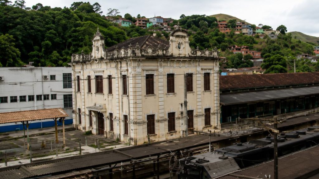 Edifício histórico de dois pavimentos com detalhes ornamentais em tons de bege ao lado de plataformas e trilhos de trem.